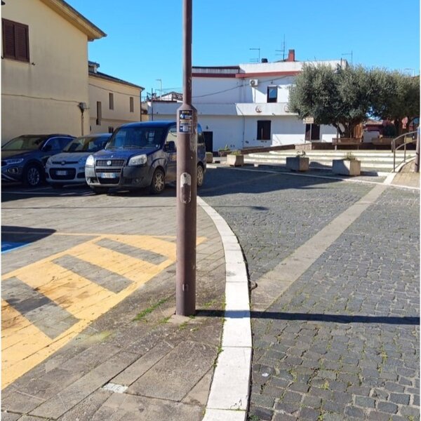 Piazza con parcheggio, lampione e edificio bianco sullo sfondo.