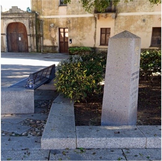 Panchina e monumento in pietra in una piazza con edificio storico sullo sfondo