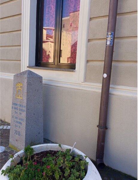 Lapide commemorativa in pietra vicino a una finestra con fiori in vaso.