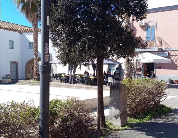 Piazza soleggiata con alberi, panchine e persone sedute al bar all'aperto.