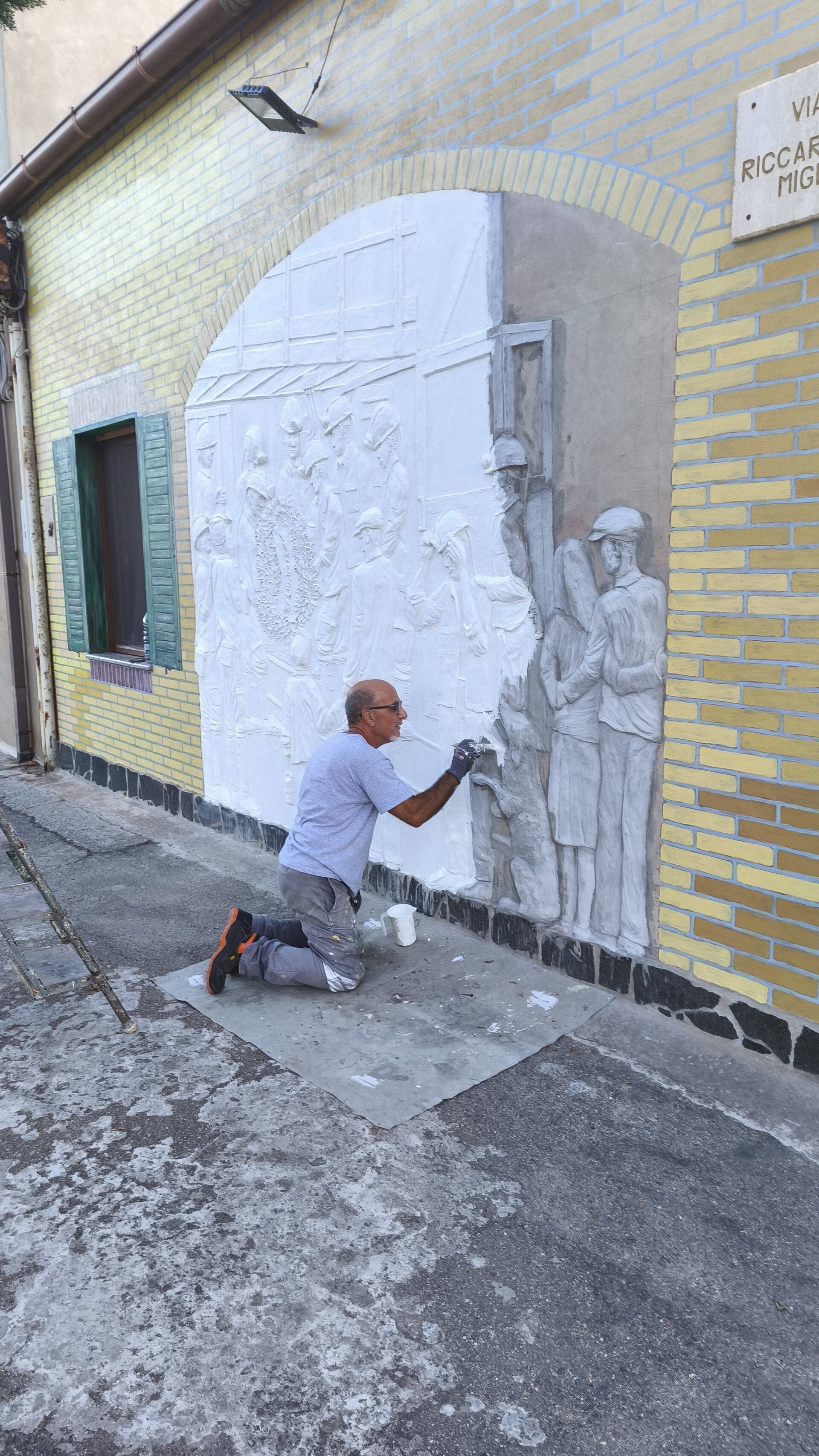 Artista dipinge un rilievo bianco su un muro esterno di mattoni gialli.