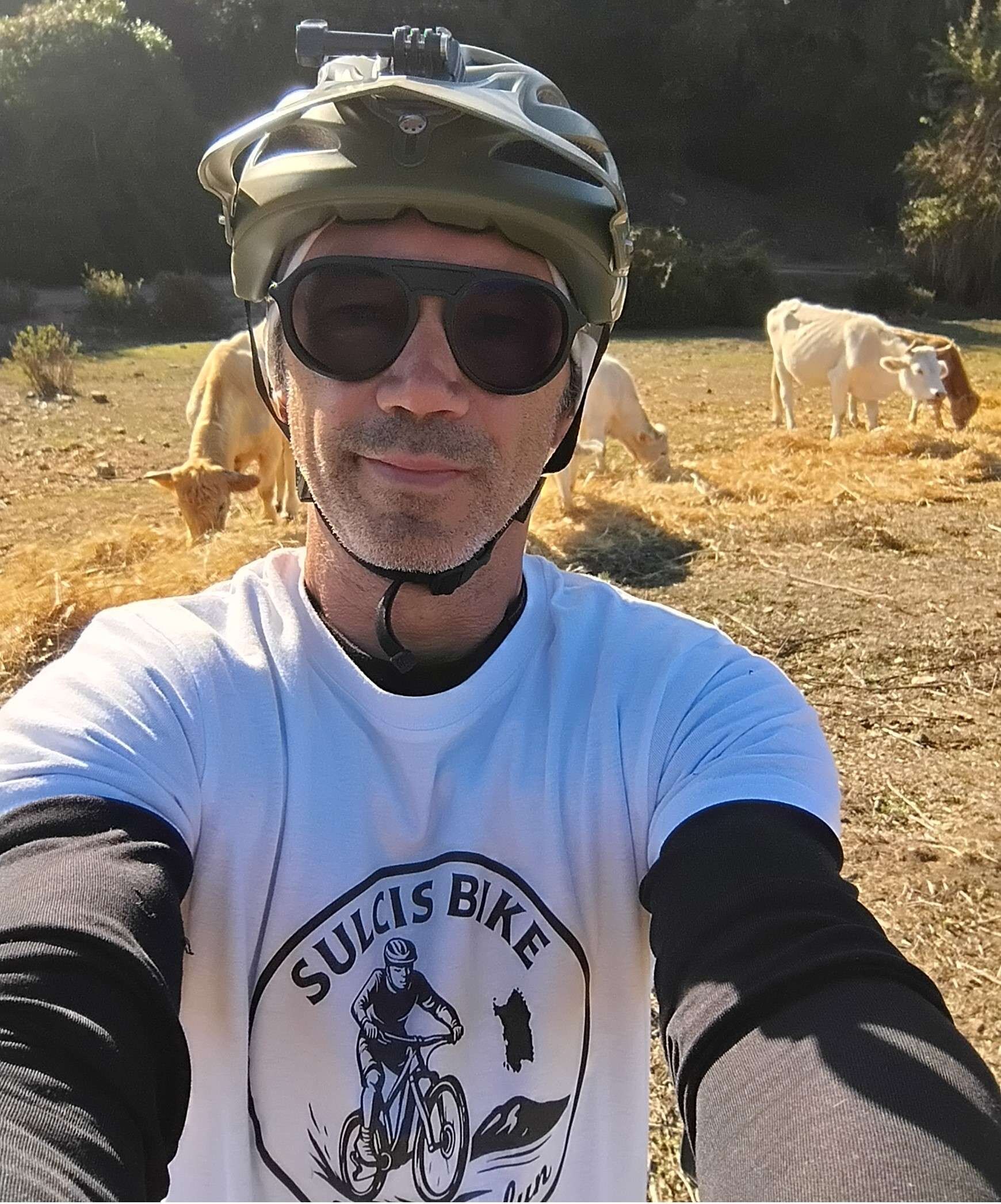 Man wearing helmet and sunglasses takes selfie in countryside with cows in background
