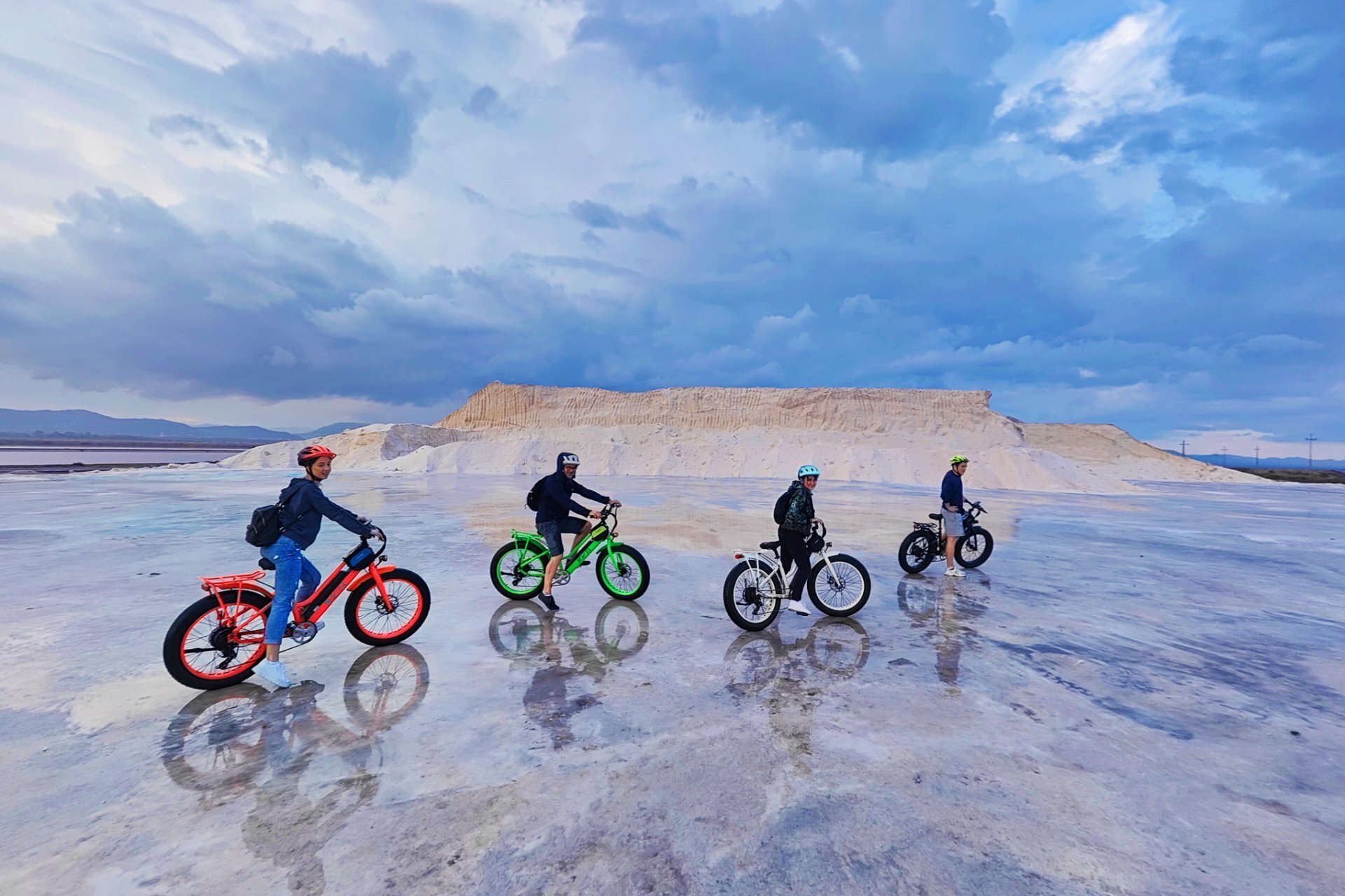 Four people riding bikes on reflective ground with cloudy sky and mountain in background.