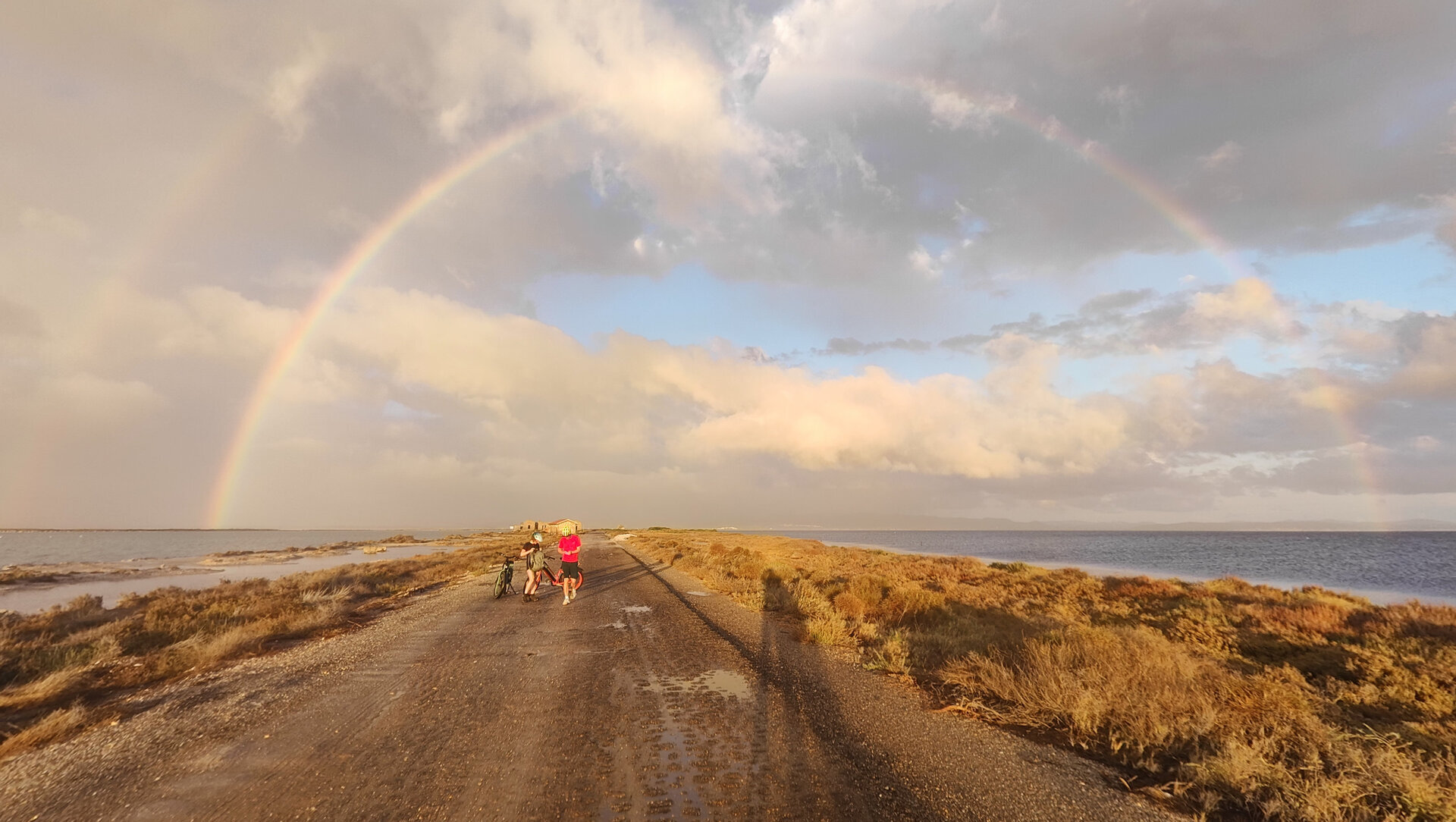 Two people walking on a dirt road near the sea under a cloudy sky.
