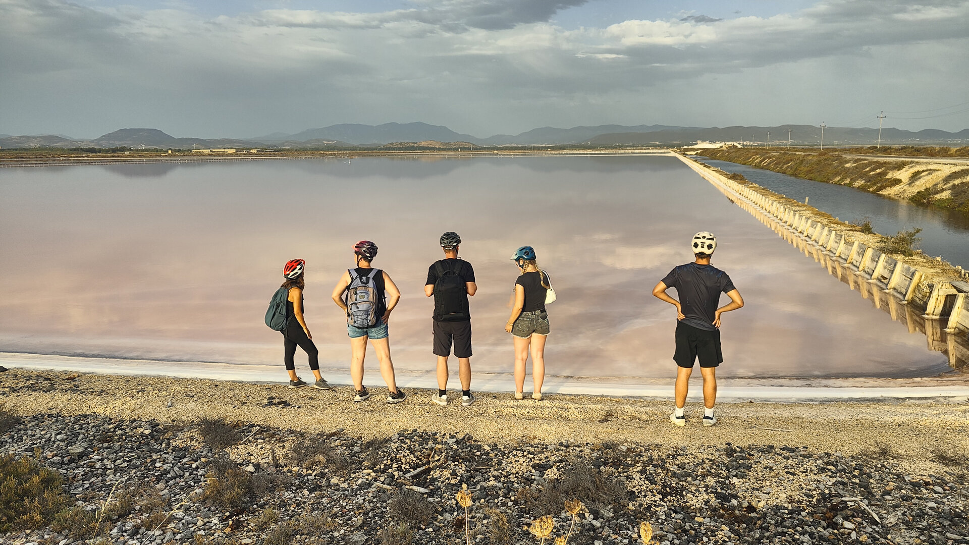 Five people wearing helmets look at a reflective salt lake with mountains in the background.