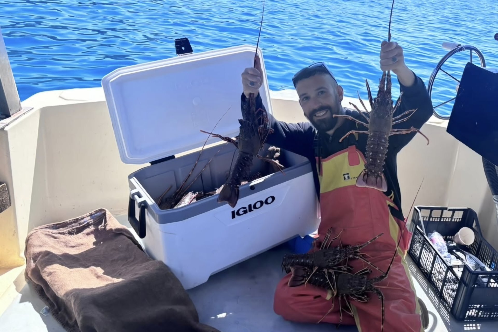 Uomo su barca mostra due grandi aragoste pescate, con ghiacciaia aperta piena di aragoste.