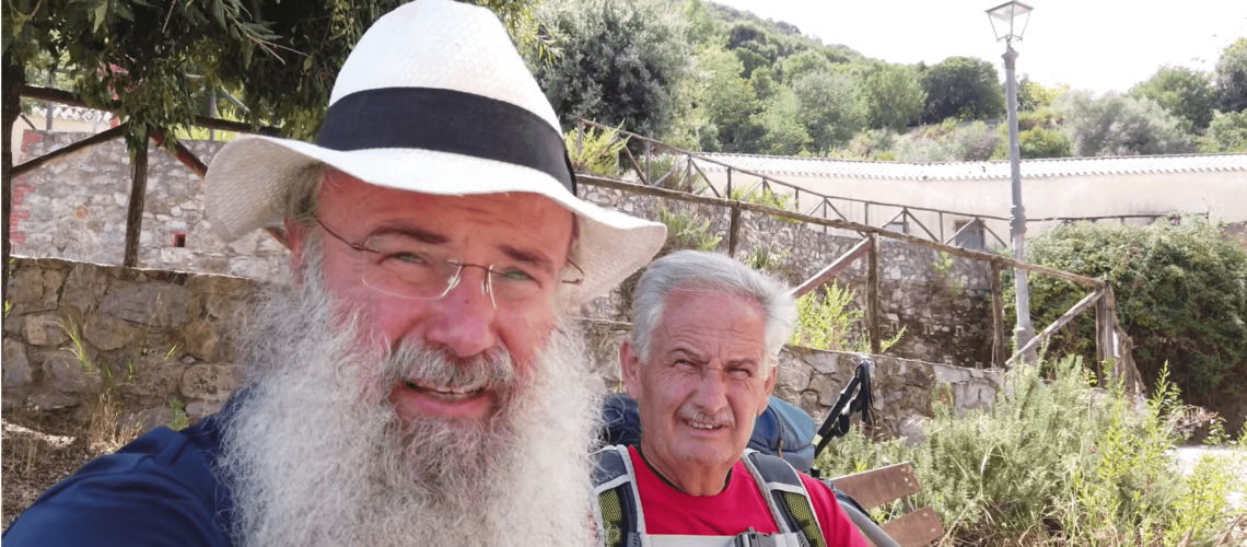 Two elderly men with backpacks in a sunny natural outdoor setting.