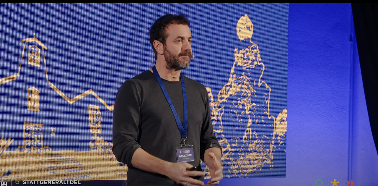 Man with headset microphone speaking on stage in front of golden graphic screen