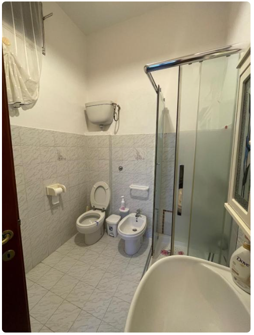Bagno con wc, bidet, doccia e lavabo in piastrelle bianche.