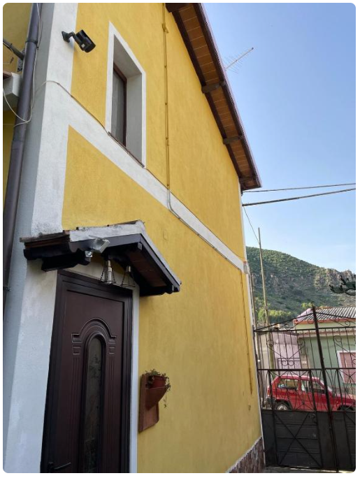 Facciata gialla di una casa con porta marrone e finestra, vista su collina e auto rossa.