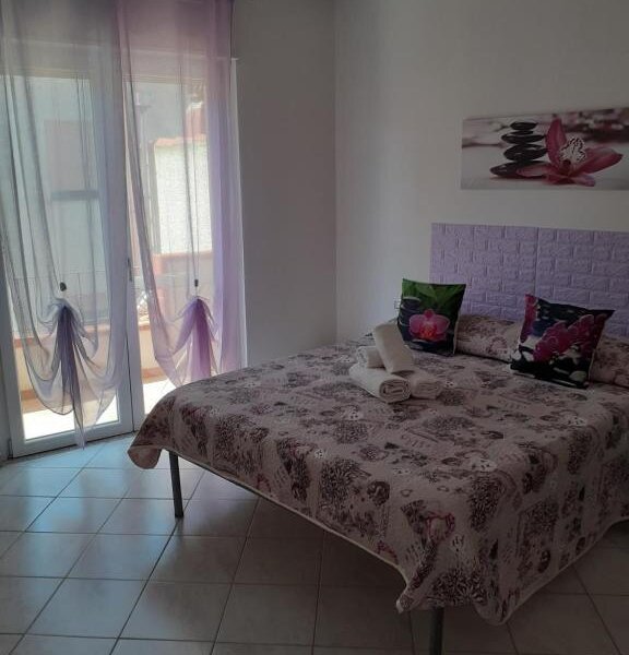 Camera da letto con letto matrimoniale, cuscini floreali e tende viola trasparenti