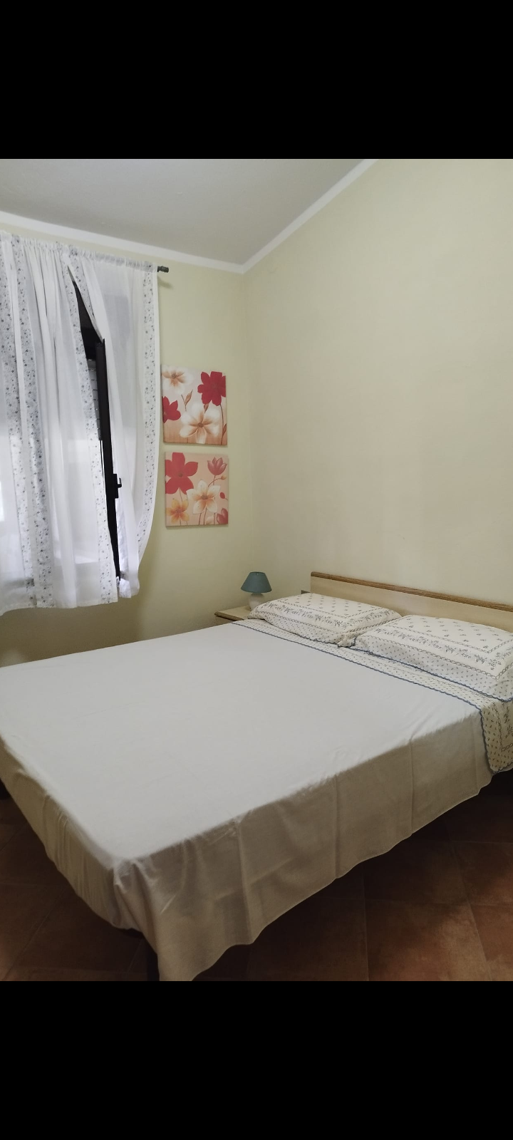 Camera da letto con letto matrimoniale, tende bianche e quadri floreali alle pareti.
