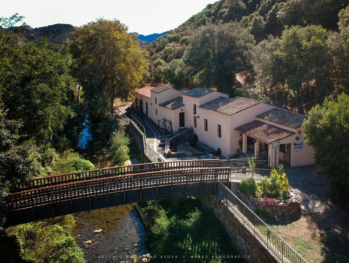 Museo etnografico vicino a un fiume con ponte pedonale e vegetazione rigogliosa