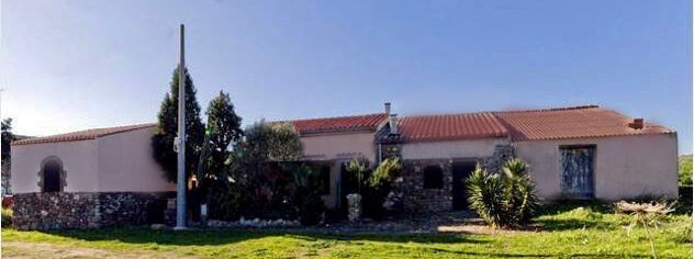 Casa rurale con tetto rosso, muro in pietra e vegetazione davanti.