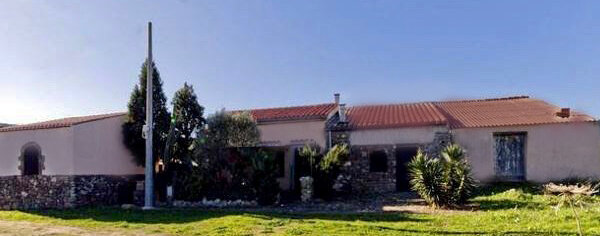 Casa rurale con tetto rosso, muro in pietra e vegetazione davanti.