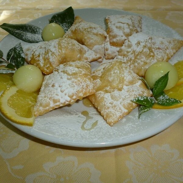 Piatto con ravioli dolci spolverati di zucchero a velo e decorazioni di agrumi e foglie verdi.